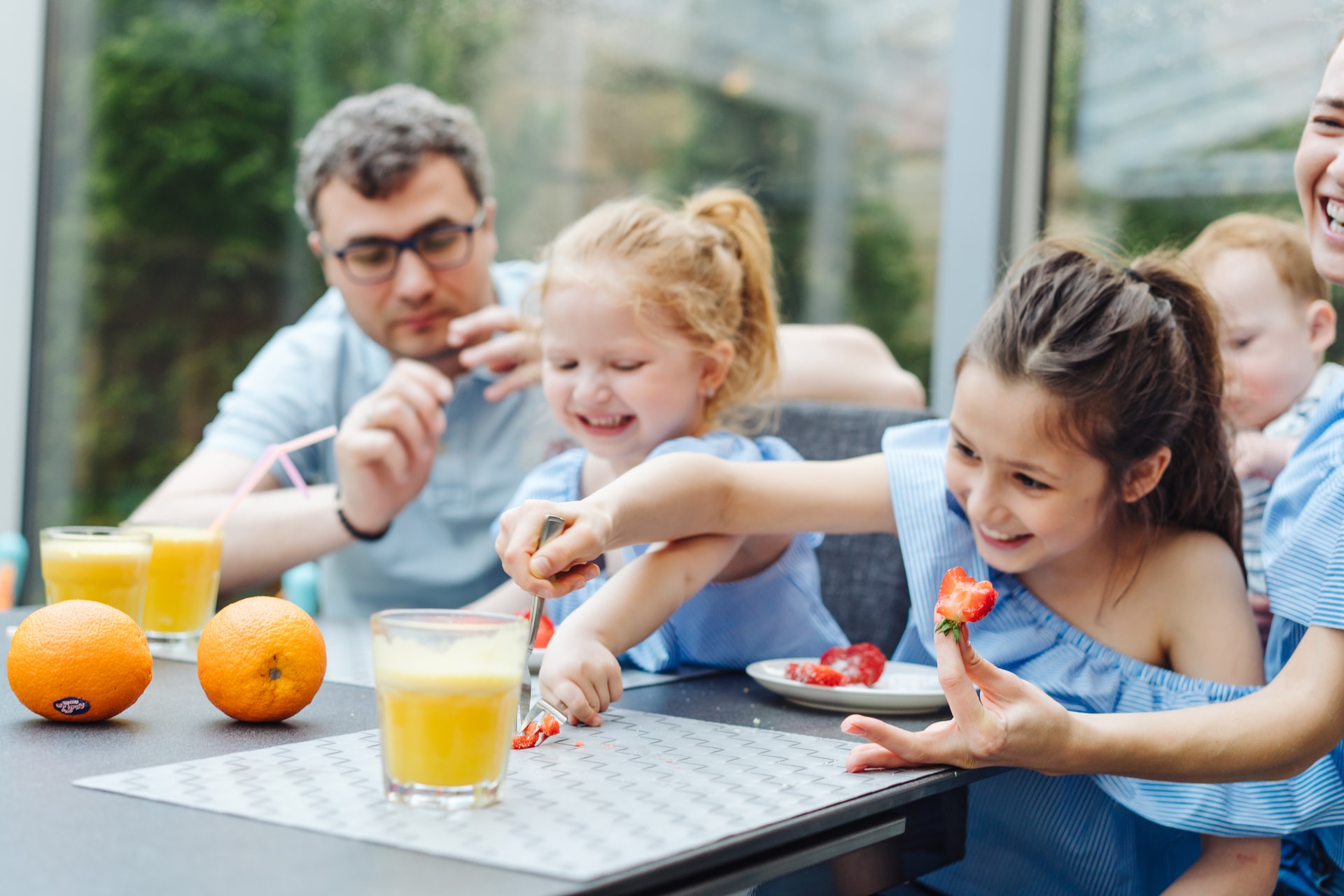 A happy family enjoys a fresh fruit breakfast, with two girls preparing strawberries and an adult overseeing, surrounded by orange juice and whole oranges.