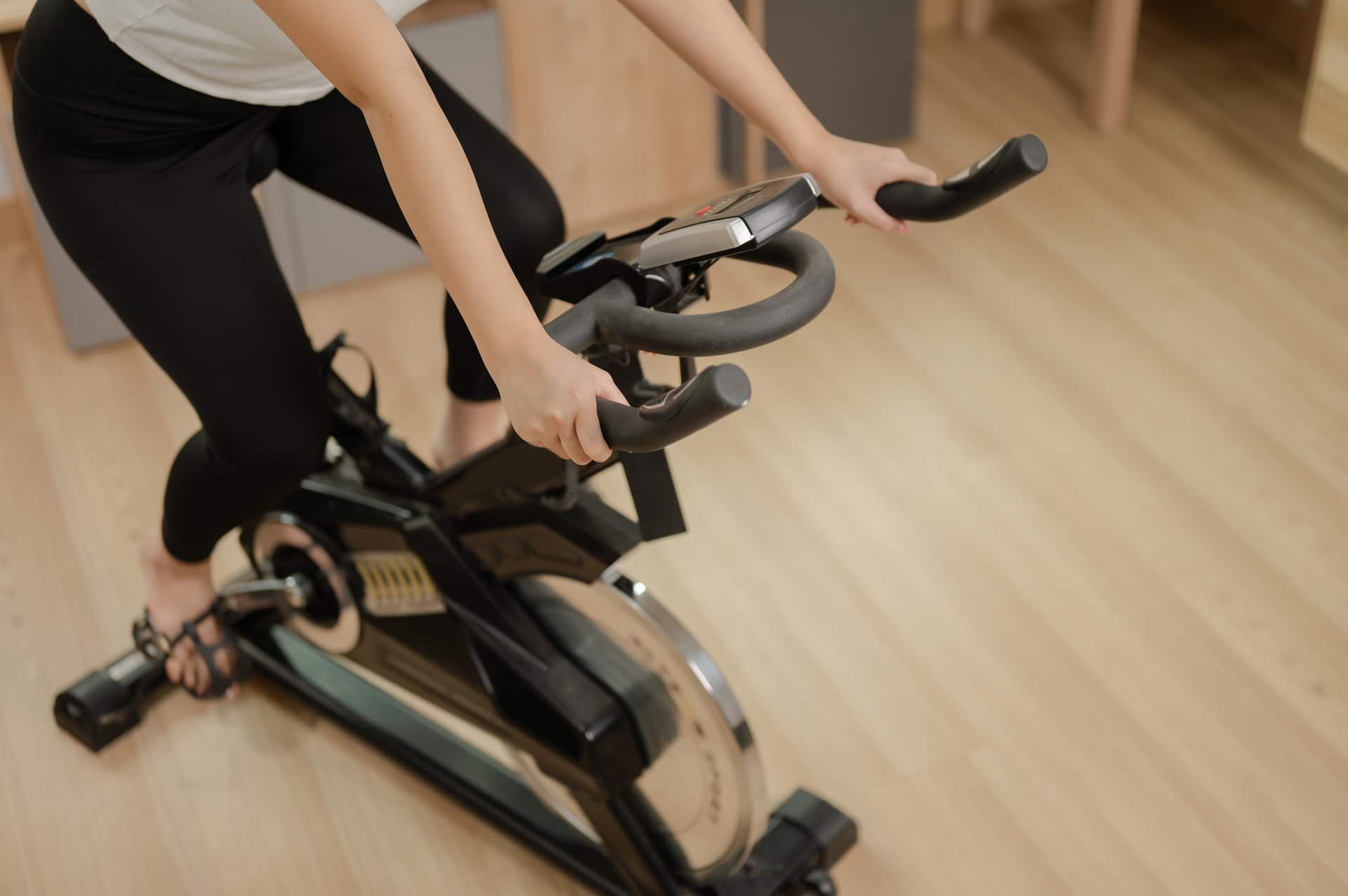 The image shows a woman in sportswear using a stationary bike for indoor cardio, with her hands gripping the handlebars. The room has a wooden floor