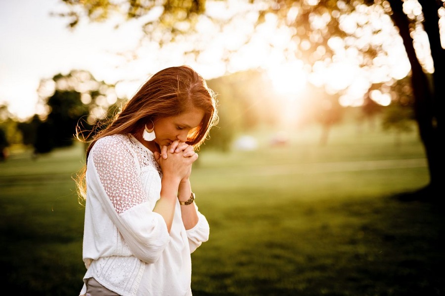 Woman folding her hands in prayer and looking down. Greenery is in the background.