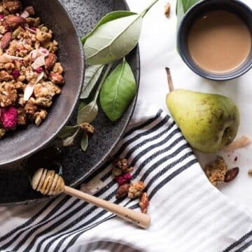 Granola with pears in a dark ceramic bowl with herbs and honey on a plate underneath it