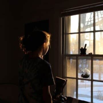 Girl standing in the dark looking out the window with sun streaming in