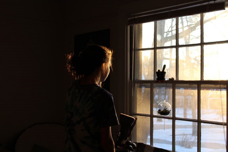 Girl standing in the dark looking out the window with sun streaming in