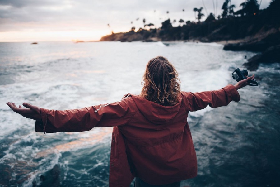 Woman in a red raincoat standing on the shore of a beach with arms outstretched