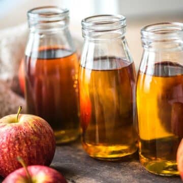 Apple juice in three glass containers surrounded by red and yellow apples and a sweater