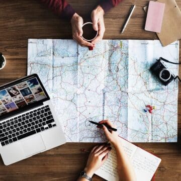 Two people's arms pointing to a map with a camera, succulents, laptop on a wooden table